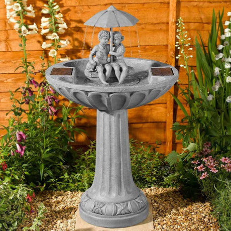 A gray stone-style garden bird bath fountain with a pedestal base, a shallow round basin, a small sculpted figure sitting under a rain umbrella, and solar panel covers on the sides, set outdoors in front of a wooden fence with plants and flowers.