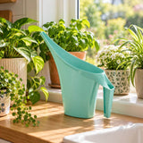A mint green plastic watering can with a long spout and integrated handle, set on a wooden windowsill surrounded by potted green plants in a bright, sunny indoor garden setting.
