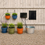 Solar-powered drip irrigation system with a black solar panel, electronic controller, and black tubing watering five potted plants against a light wood fence and gravel ground.