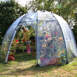 A large green dome greenhouse made of PVC with a fibreglass structure, set up outdoors with plants inside and a person standing outside.