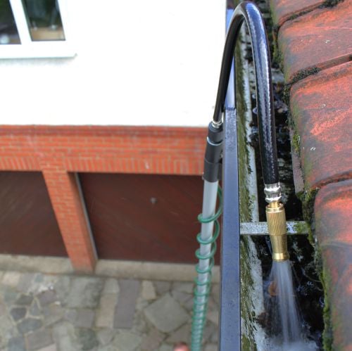 A flexible gutter cleaner with a green hose attached to a brass nozzle, shown in use cleaning a gutter.