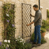 Man adjusting a decorative willow trellis against a stone wall with flowers and plants.