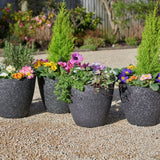 Four grey plant pots with granite effect, filled with colourful flowers on a gravel surface, garden background.