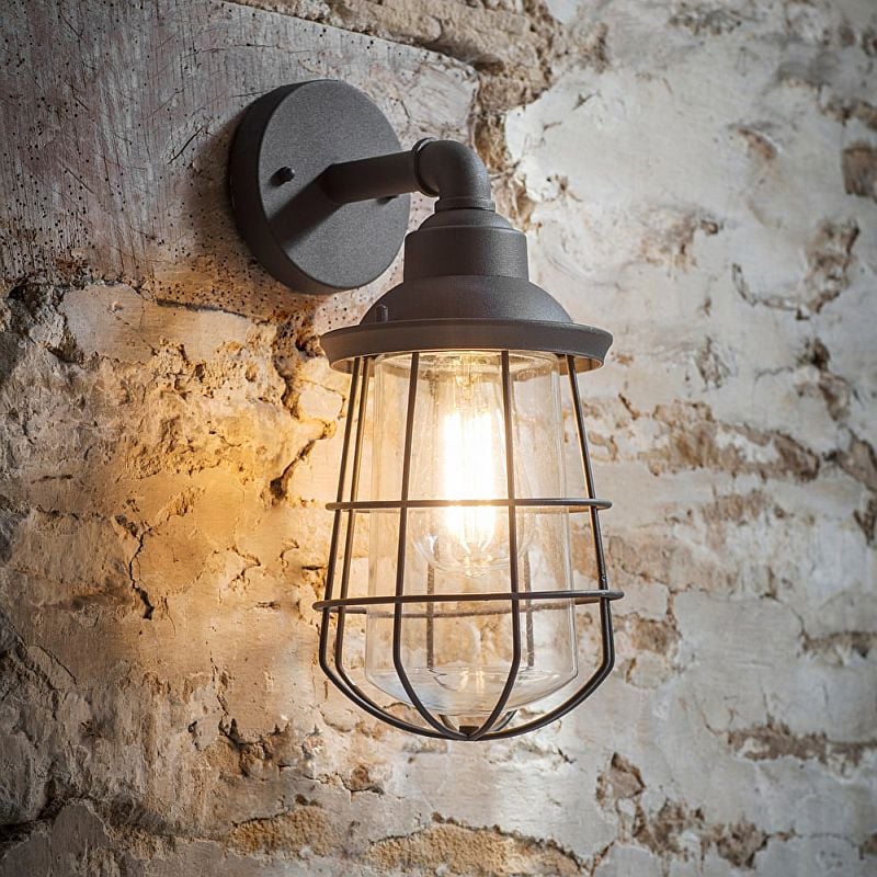 An outdoor wall-mounted light fixture with a charcoal matt finish and a glass cage, mounted on a brick wall.