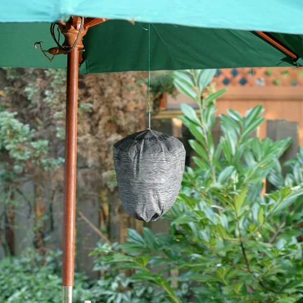 A gray, fabric wasp repeller hanging from a green umbrella, surrounded by greenery in the background.