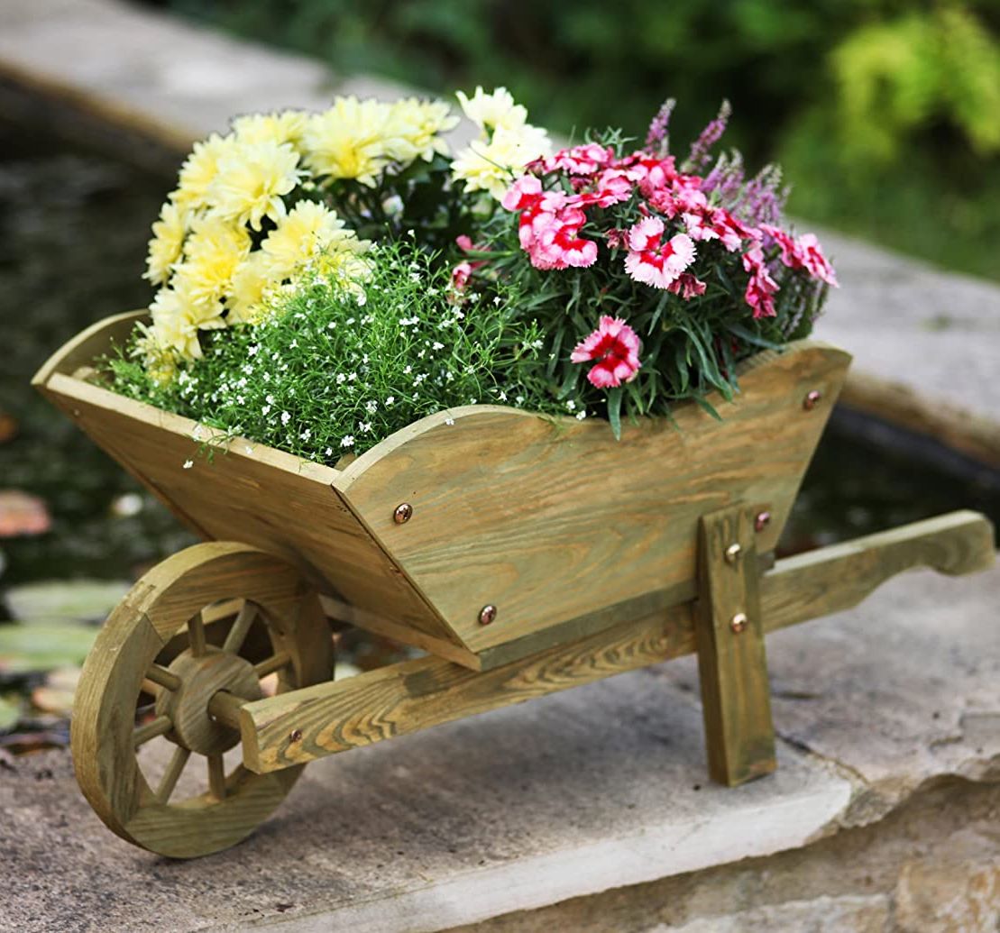 A wooden wheelbarrow planter filled with various flowers, placed on a stone surface with a brick wall in the background.