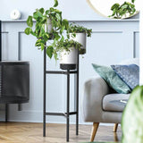 A white plant pot stand with three levels, featuring a sturdy industrial metal frame, placed in a room with various plants and a grey sofa.
