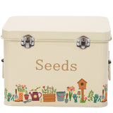 Cream-colored metal seed storage tin with 'Seeds' printed on the front, decorated with a colorful border of gardening tools and plants along the bottom edge.