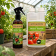 Two garden products from gardening-naturally displayed on a wooden table inside a greenhouse: a brown spray bottle labeled Whitefly Spray and a white jug labeled Whitefly Treatment, both with tomato graphics on the labels.