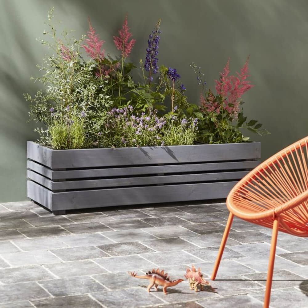 Gray wooden planter with flowers on a stone patio with an orange chair and dinosaur toys.