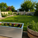 A sunny backyard garden scene with a green lawn, wooden fence, trees, a small patio set, and a soccer goal. In the foreground, a large rectangular metal planter box with flowers sits on the left, and part of a wooden barrel planter is visible on the right.