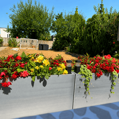 Gray rectangular zinc planters filled with red and yellow flowers in a backyard setting with trees and a wooden fence in the background.